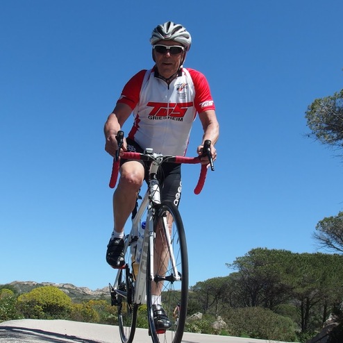 Radfahrer in sportlicher Kleidung fährt auf einer Landstraße, umgeben von Bäumen und blauem Himmel.