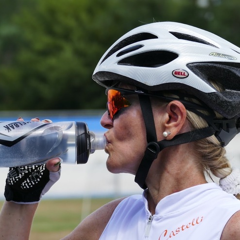 Frau mit Fahrradhelm trinkt aus einer transparenten Wasserflasche auf einem Sportgelände.