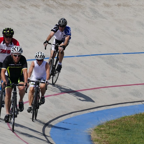 Fünf Radfahrer fahren auf einer velodromartigen Bahn mit farbigen Markierungen. Grüne Wiese am Rand sichtbar.