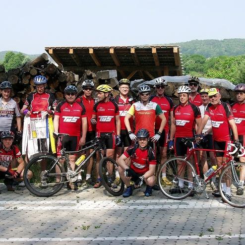 Gruppe von Radfahrern in roten Trikots vor einem Holzstapel, einige mit Fahrrädern, umgeben von grüner Landschaft.