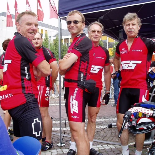 Sechs Radfahrer in roten Trikots stehen zusammen, lächeln und bereiten sich auf ein Rennen vor.