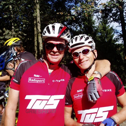 Zwei Radfahrer in roten Trikots posieren lächelnd im Wald, umgeben von anderen Radfahrern und Fahrrädern.