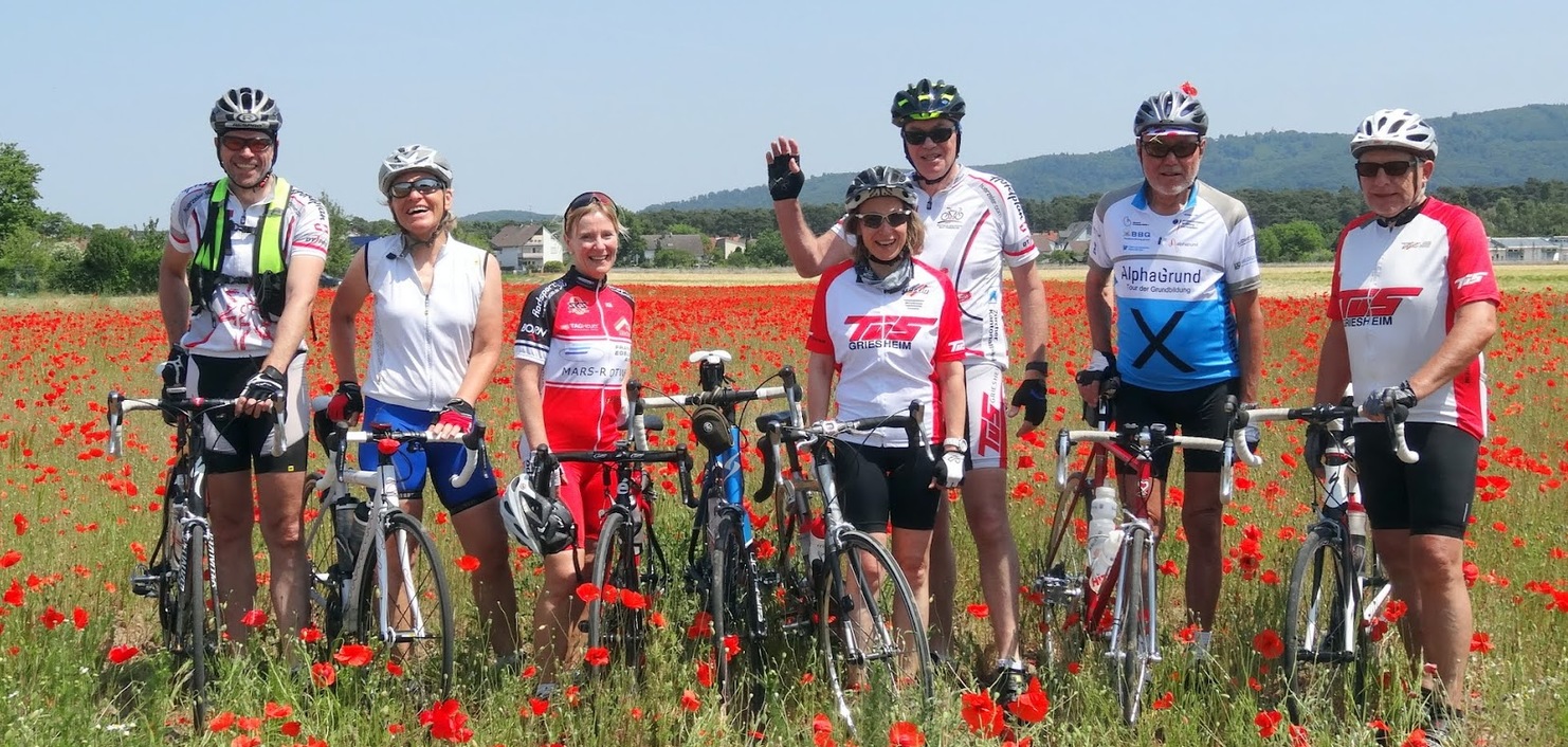 Gruppe von Radfahrern steht lächelnd in einem Mohnfeld, umgeben von leuchtend roten Blumen und blauen Himmel.