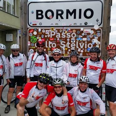 Gruppe von Radfahrern in Trikots vor einem Schild in Bormio, Passo Stelvio, umgeben von Aufklebern und Bergen.