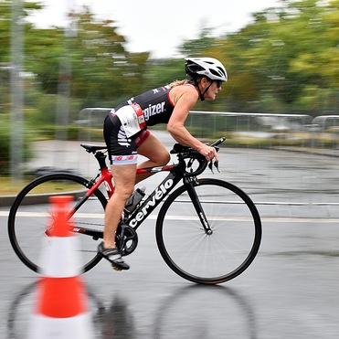 Sportlerin in Radleroutfit fährt schnell auf einem Rennrad mit regennasser Straße im Hintergrund. Verkehrshütchen in der Nähe.