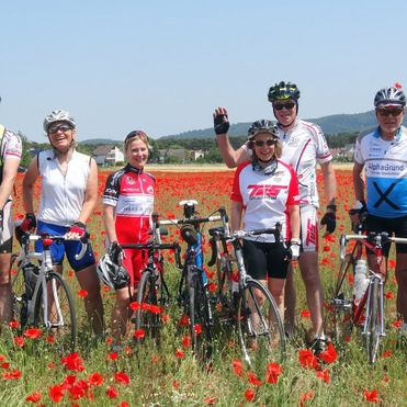 Gruppe von Radfahrern steht lächelnd in einem Mohnfeld, umgeben von leuchtend roten Blumen und blauen Himmel.