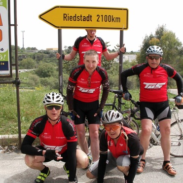 Gruppe von fünf Radfahrern in roten Trikots posiert vor einem „Riedstadt 2100km“-Schild an einer Landstraße.