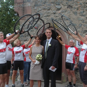 Brautpaar steht vor einem Eingang, während Radfahrer mit Fahrradrädern jubelnd anstoßen.