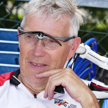 Mann mit Brille und Sportkleidung sitzt nachdenklich auf einem Stuhl, Fahrrad im Hintergrund.