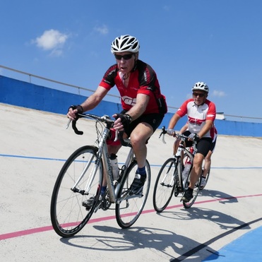 Zwei Radfahrer in roten und weißen Trikots fahren auf einer Radrennbahn mit blauer und grauer Oberfläche.