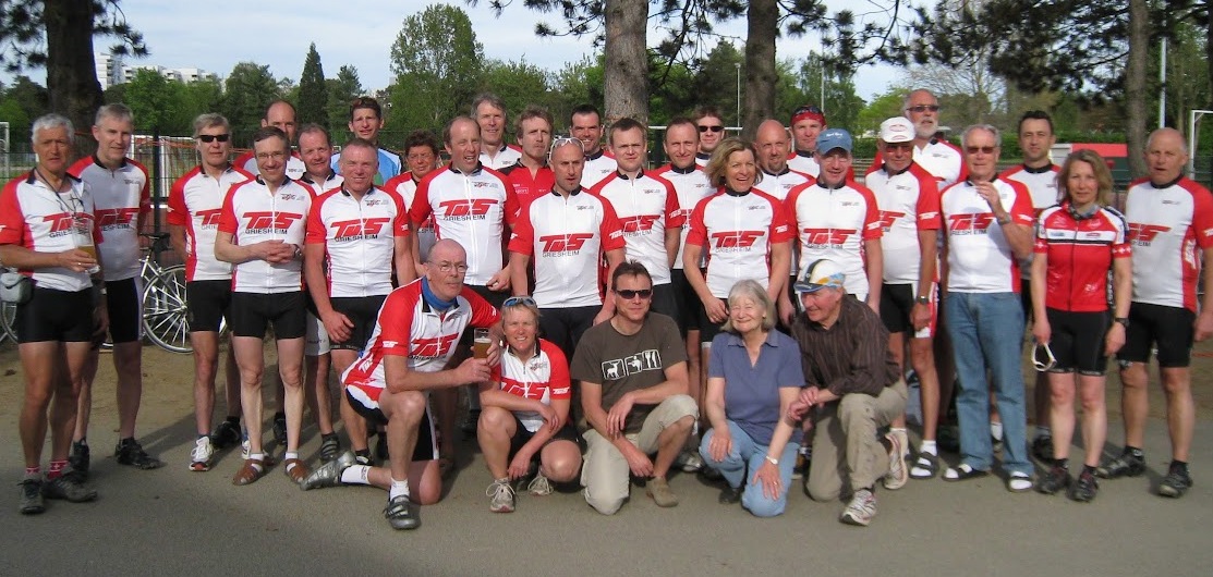 Gruppenfoto von Radfahrern in roten und wei&szlig;en Trikots, stehend vor einer Sportanlage unter B&auml;umen.