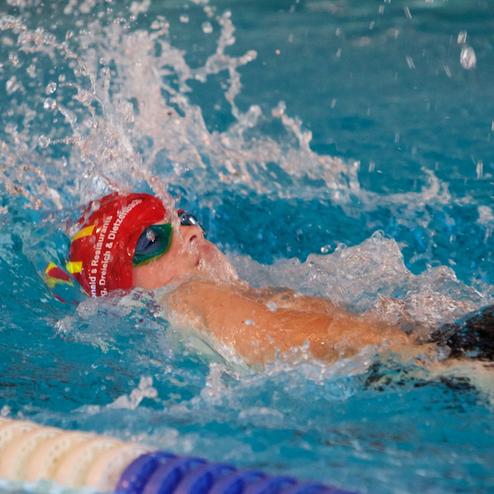 Schwimmerin mit roter Badekappe und Sonnenbrille gleitet durch das Wasser, umgeben von Spritzwasser.