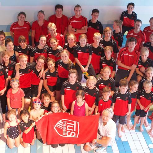Gruppenshooting von Schwimmern in roten Trikots, zusammen mit Trainerin, vor einem Schwimmbecken.