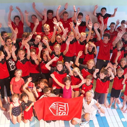 Gruppenfoto von Schwimmern in roten T-Shirts, die im Schwimmbad die Hände heben und ein Banner halten.