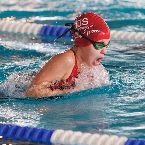 Junge Schwimmerin im roten Badeanzug und Badekappe beim Wettkampf im Wasser, spritzt Wasser beim Kraulen.
