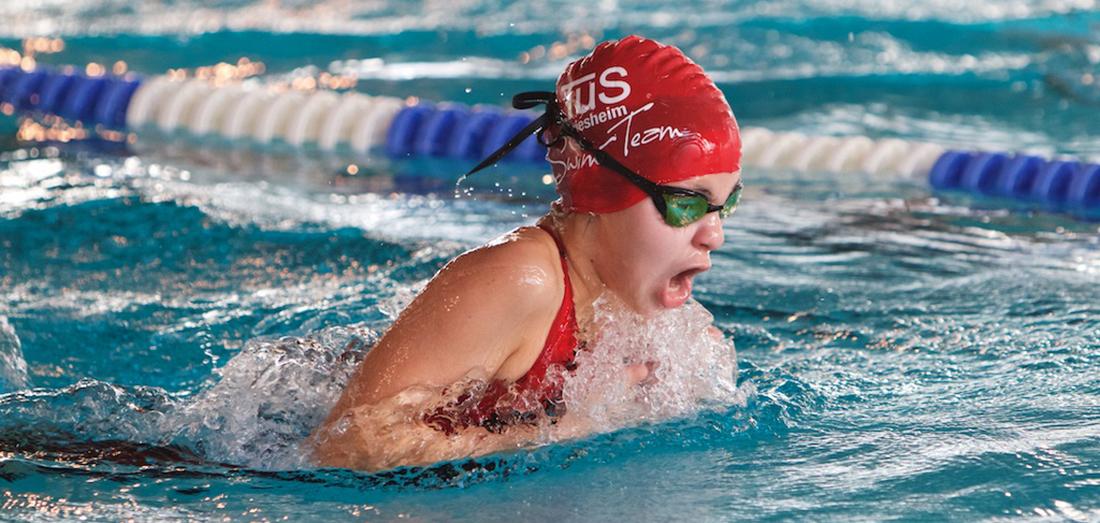 Junge Schwimmerin im roten Badeanzug und Badekappe beim Wettkampf im Wasser, spritzt Wasser beim Kraulen.