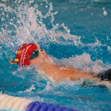 Schwimmerin mit roter Badekappe und Sonnenbrille gleitet durch das Wasser, umgeben von Spritzwasser.