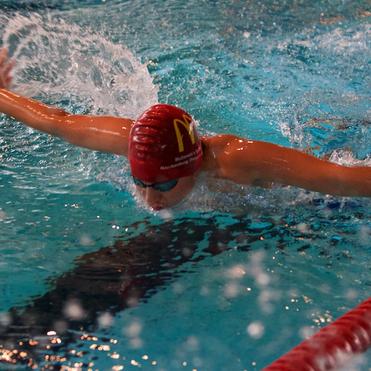 Schwimmer, der im Wasser im Schmetterlingsstil schwimmt, mit roter Schwimmhaube und Brille auf. Spritzer im Wasser sichtbar.