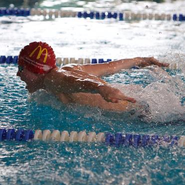 Schwimmer mit roter Badekappe im Wasser, der mit Kraultechnik einen Schlag ausführt. Spritzwasser um ihn herum.