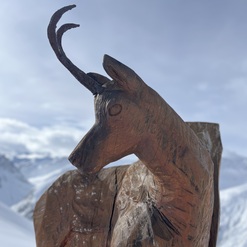 Holzskulptur eines Rehs mit sichtbaren Geweih auf schneebedecktem Hintergrund und bewölktem Himmel.