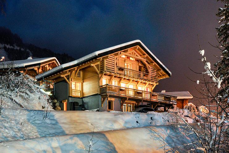 Einfamilienhaus im Schnee bei Nacht, beleuchtet mit warmen Lichtern und umgeben von verschneiten Bäumen und Bergen.