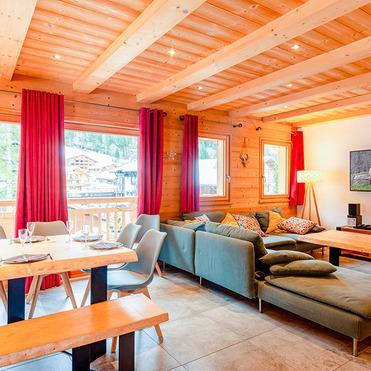 Heller Wohnraum mit Holzdecken, großen Fenstern, rotem Vorhang, Sofalandschaft und Essbereich aus Holz. Fernseher an der Wand.