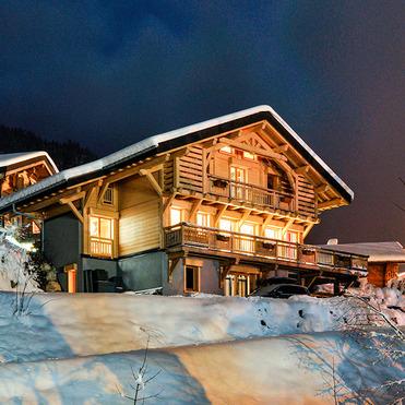 Einfamilienhaus im Schnee bei Nacht, beleuchtet mit warmen Lichtern und umgeben von verschneiten Bäumen und Bergen.