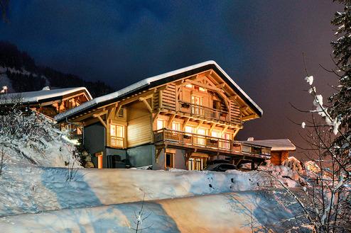Einfamilienhaus im Schnee bei Nacht, beleuchtet mit warmen Lichtern und umgeben von verschneiten Bäumen und Bergen.