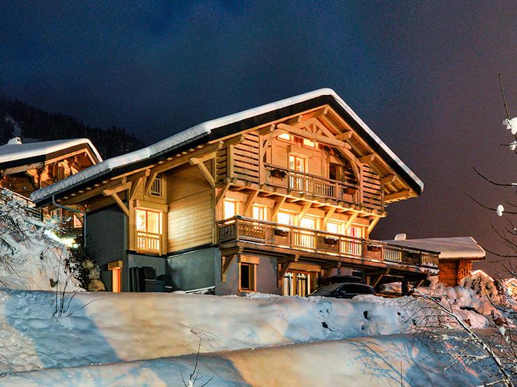 Einfamilienhaus im Schnee bei Nacht, beleuchtet mit warmen Lichtern und umgeben von verschneiten Bäumen und Bergen.