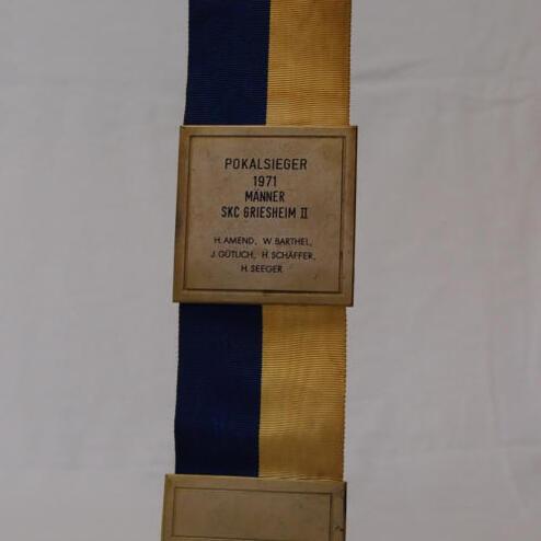 Pokal in gold, blau-gelber Stoffband mit Inschrift "Pokalsieger 1971 Männer SKC Griesheim II" und Namen.