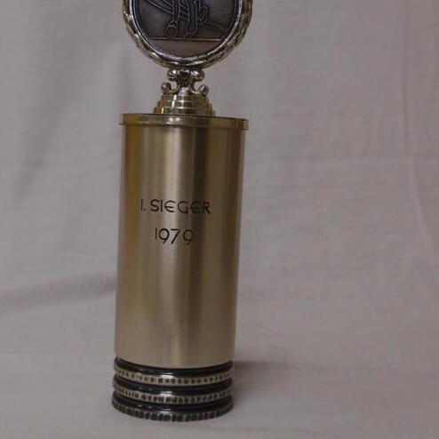 Pokaltrophäe mit der Aufschrift „I. SIEGER 1979“ und einer Medaille an der Spitze, auf einem weißen Hintergrund.