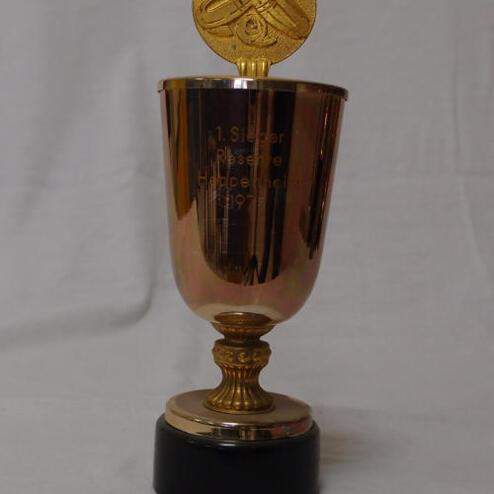 Goldfarbener Pokal mit Gravur „1. Sieger Plazierung 1997“ auf einem schwarzen Sockel.