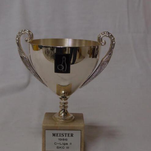 Silberner Pokal mit zwei Griffen, graviert mit "Meister 1986 C-Liga II SKC III" auf einem marmorierten Sockel.