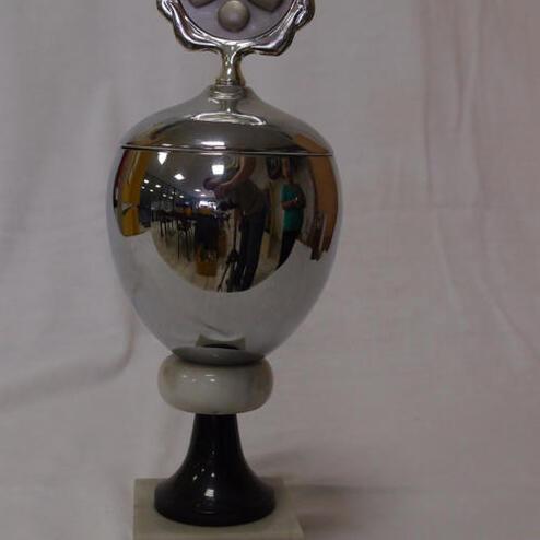 Schimmernder Sportpokal auf schwarzem Sockel mit gravierter Inschrift und Zinnmedaille mit Sportmotiven oben.