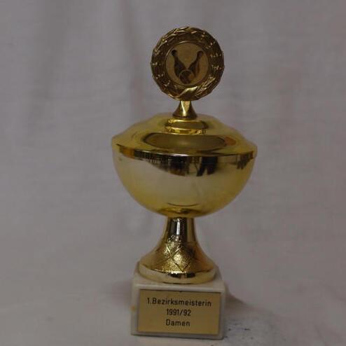 Goldener Pokal mit der Inschrift „1. Bezirksmeisterin 1991/92 Damen“ auf einem weißen Sockel.