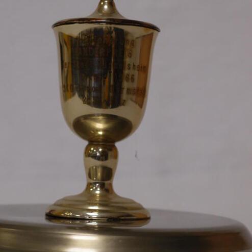 Goldener Pokal auf einem silbernen Untersetzer, mit eingravierter Beschriftung, leicht schräg abgebildet.