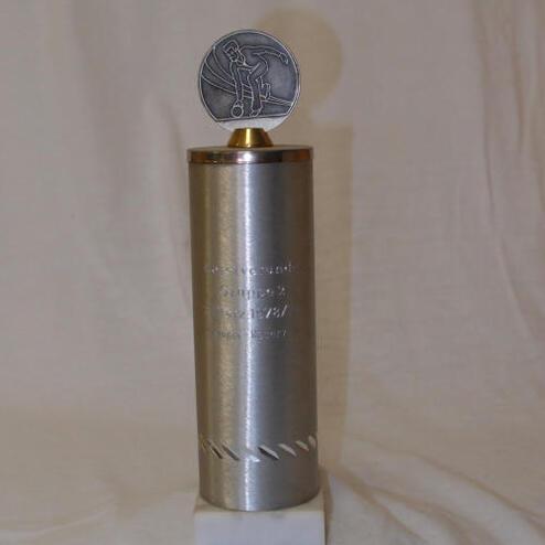Silberner Pokal mit kreisförmiger Medaille oben, auf weißem Sockel, mit eingraviertem Text.