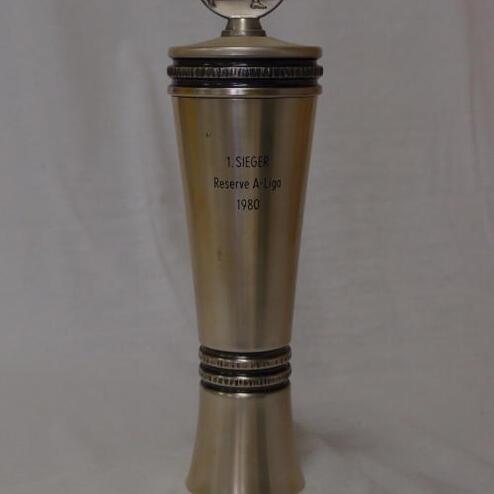 Pokale mit Aufschrift „1. Sieger Reserve A-Liga 1980“ und einer Sportlerfigur als Deckel auf grauem Hintergrund.