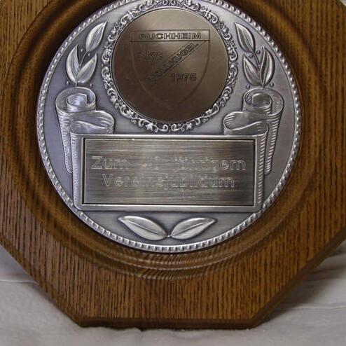 Plakette aus Holz mit silberner Medaille, verzierte Ränder und Inschrift „Zum Jubiläum Vereinsgründung“.