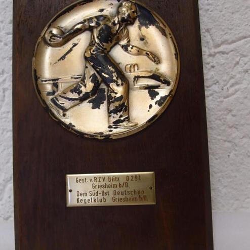 Plakette aus Holz mit einer goldenen Medaille, die einen Kegler in Aktion darstellt, mit eingeprägtem Text.