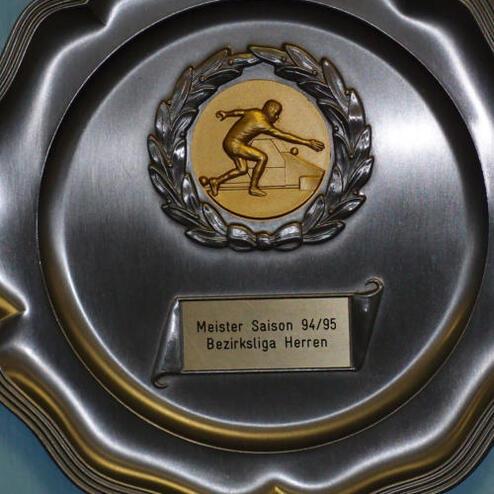 Silbernes Meisterschaftsschild mit einer goldenen Medaille und der Aufschrift „Meister Saison 94/95 Bezirksliga Herren“.
