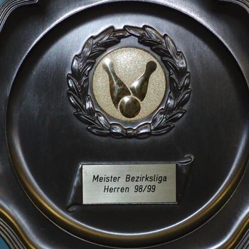 Edelmetallpreis mit Wappen und Inschrift "Meister Bezirksliga Herren 98/99" auf blauer Wand.