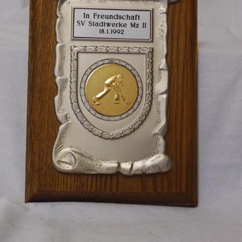 Ehrenplaque aus Holz mit silbernem Schild und goldener Medaille, beschriftet mit „In Freundschaft SV Stadtwerke Mz II 18.1.1992“.