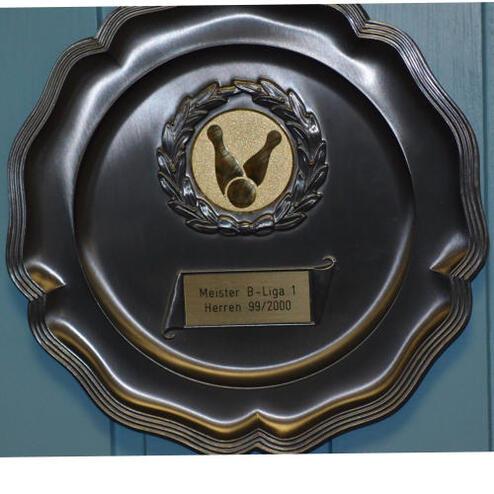 Plakette mit goldener Auszeichnung für Meisterschaft in B-Liga 1 Herren 99/2000, im Hintergrund eine blaue Wand.