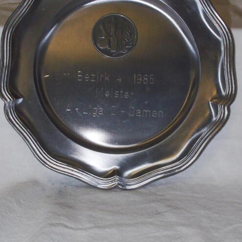 Silberner Pokal mit geprägter Aufschrift "Bezirk 1985 Meister AK-Liga 2 - Damen" auf weißem Hintergrund.