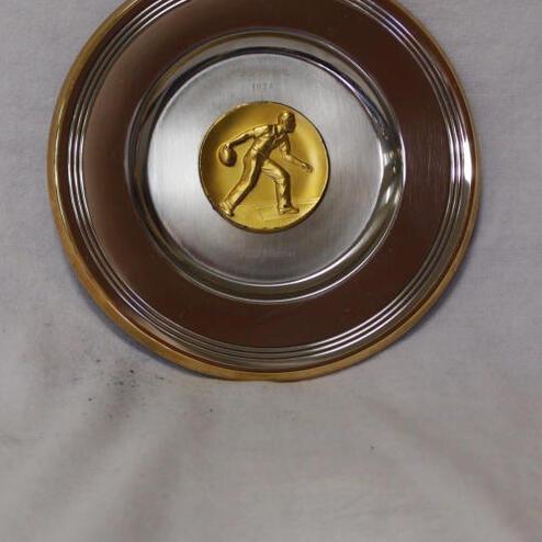 Runder Metallplakette mit goldenem Sportler-Emblem in der Mitte auf silberfarbener Fläche.