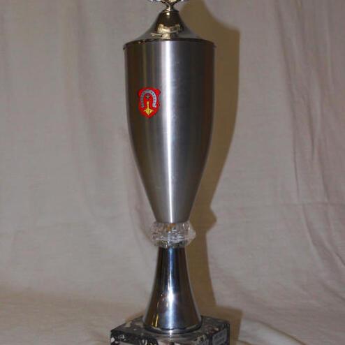 Silberner Pokal mit dekorativem Deckel, Wappen und schwarzer Basis auf hellem Hintergrund.