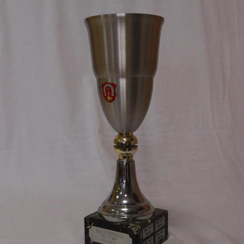 Silberne Trophäe mit goldenem Sockel und Wappen auf schwarzem Grund, steht auf einem hellen Untergrund.