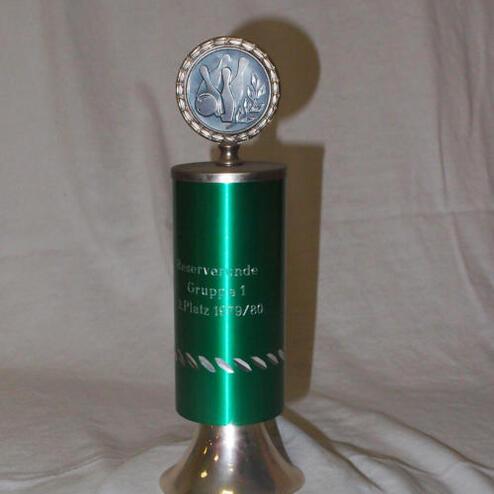 Grüner Siegerpokal mit Silbermedaille, graviert mit "1. Platz 1979/80", auf einem hellen Hintergrund.