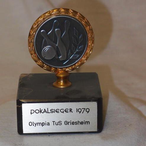 Pokal mit goldfarbener Umrandung, beschriftet mit „Pokalsieger 1979, Olympia TuS Griesheim“, auf schwarzem Sockel.